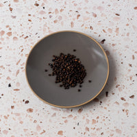 Black Peppercorns are scattered in the center of a round gray plate, displayed on a white terrazzo surface with pink and brown flecks.