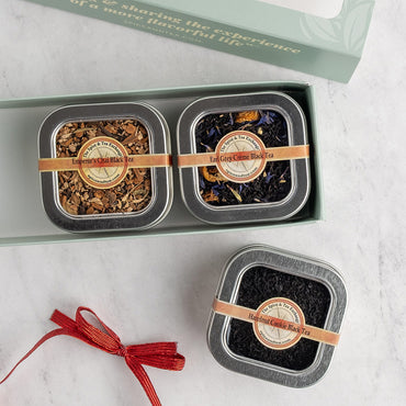 Three square metal tins of black tea arranged on a light marble surface, with one open box showing two tea blends and one tin below tied with a red ribbon. The tins have silver lids, tan bands, and round labels.