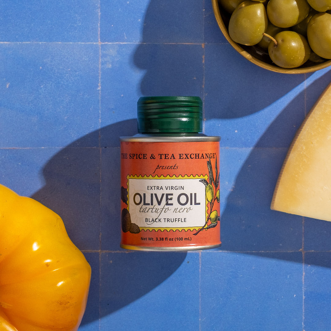 Can of  Black Truffle Extra Virgin Olive Oil - Mini with a yellow pepper and green olives on a blue tiled background