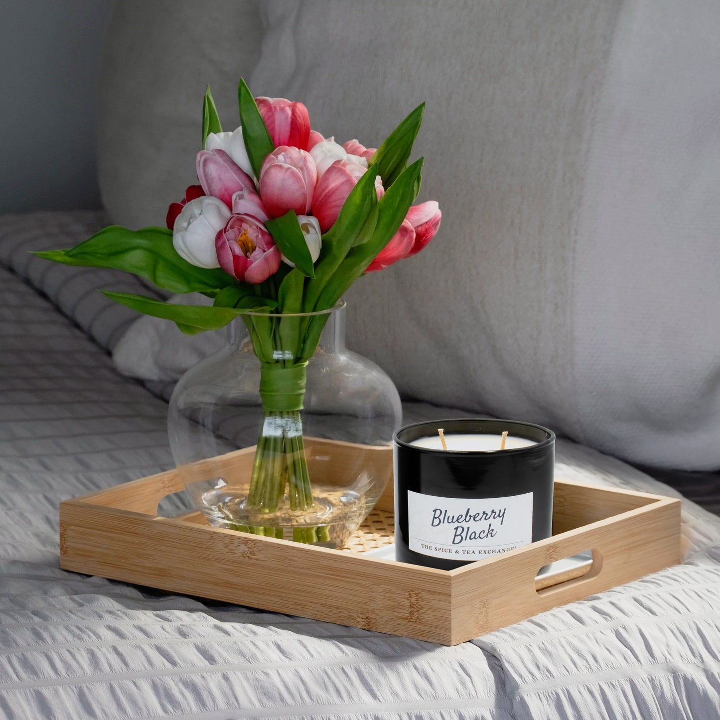Bouquet of pink and white tulips in a clear vase on a wooden tray with a black candle labeled 'Blueberry Black' on a bed.