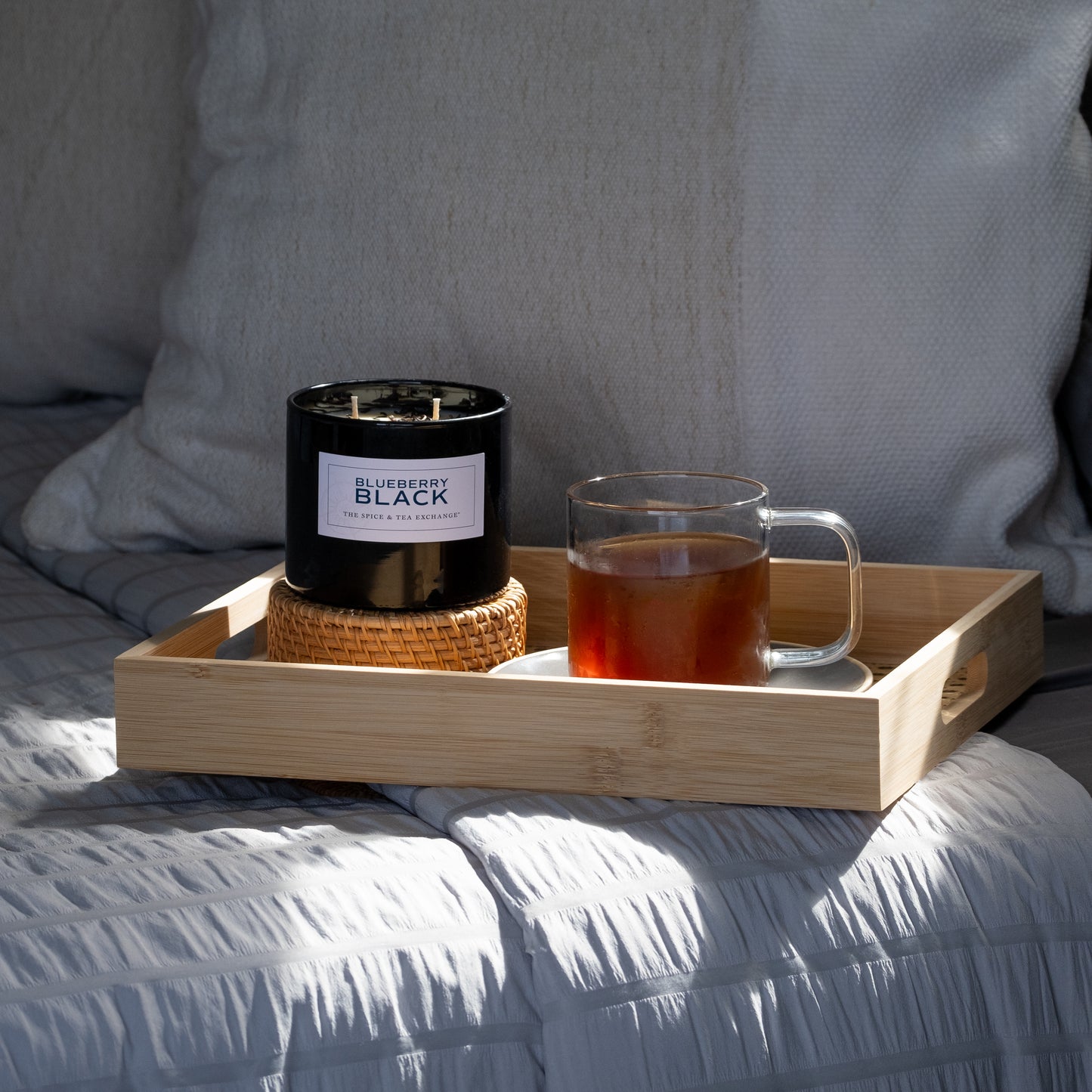 A wooden tray on the bed holds a glass mug of tea and a Blueberry Black Candle made from natural soy and essential oils, set on a wicker coaster, with pillows and a gray quilt in the background.