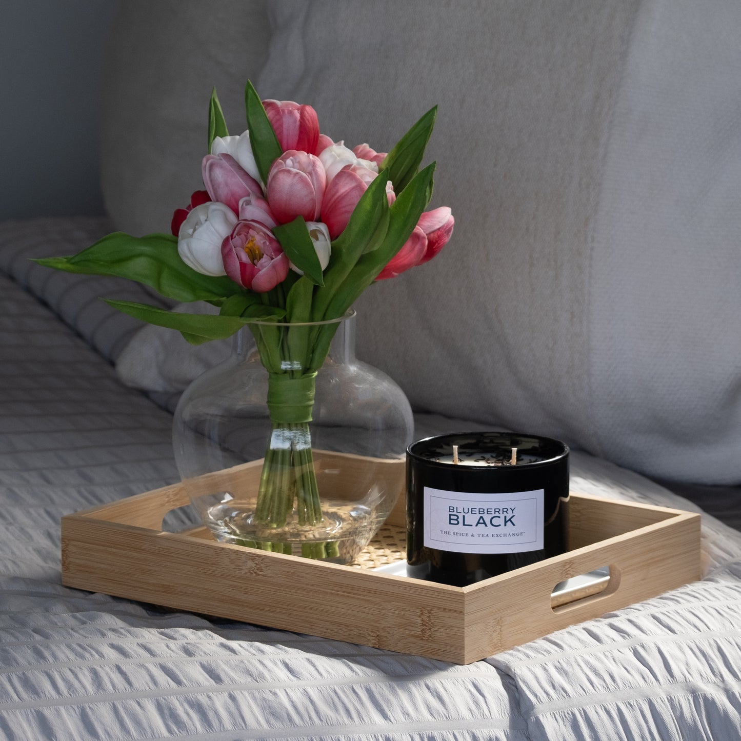 A wooden tray on the bed holds a glass vase with pink and white tulips and a Blueberry Black Candle made from natural soy and essential oils. The bed showcases light-colored, textured bedding for a cozy feel.