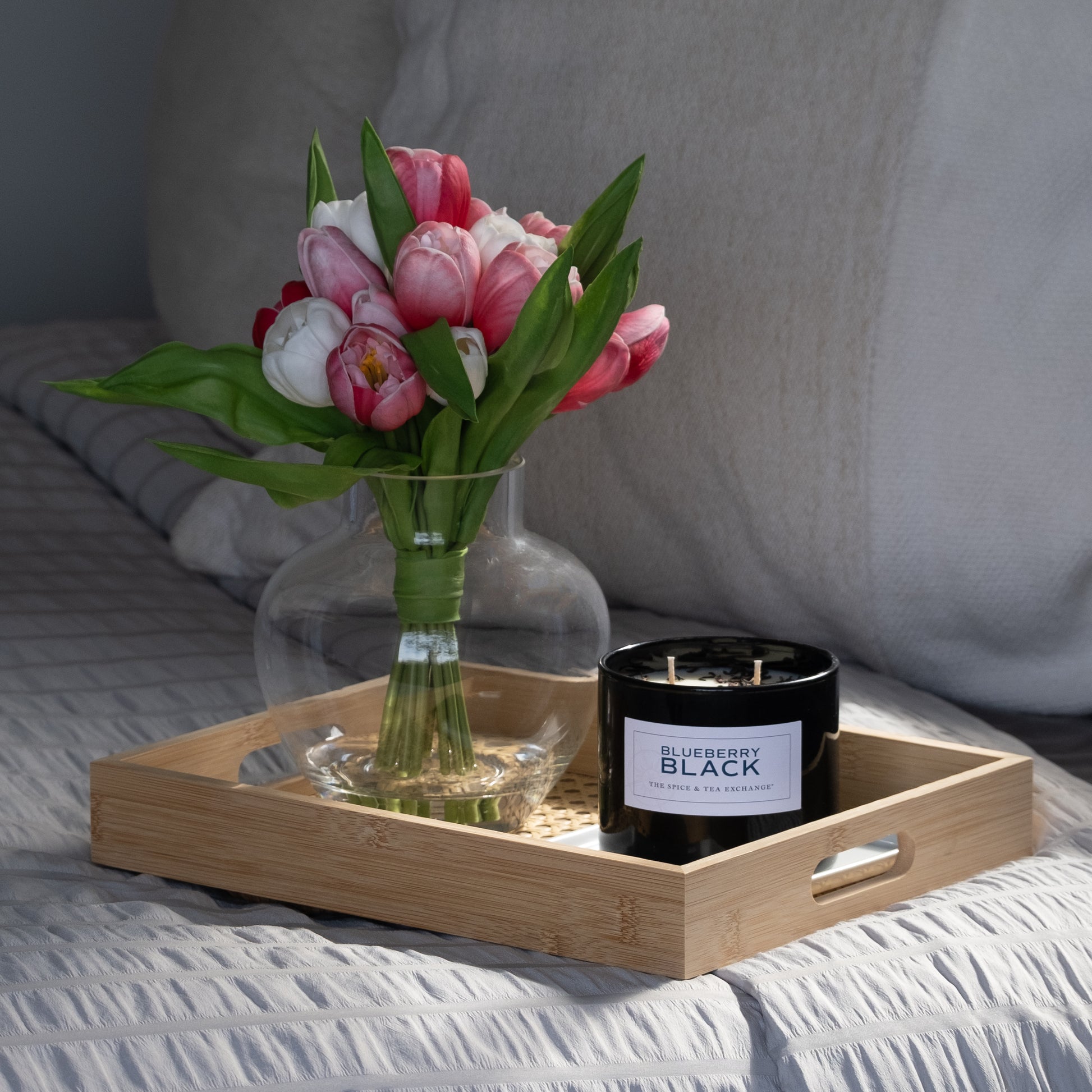 A wooden tray on the bed holds a glass vase with pink and white tulips and a Blueberry Black Candle made from natural soy and essential oils. The bed showcases light-colored, textured bedding for a cozy feel.