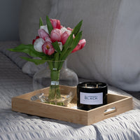 A wooden tray on the bed holds a glass vase with pink and white tulips and a Blueberry Black Candle made from natural soy and essential oils. The bed showcases light-colored, textured bedding for a cozy feel.