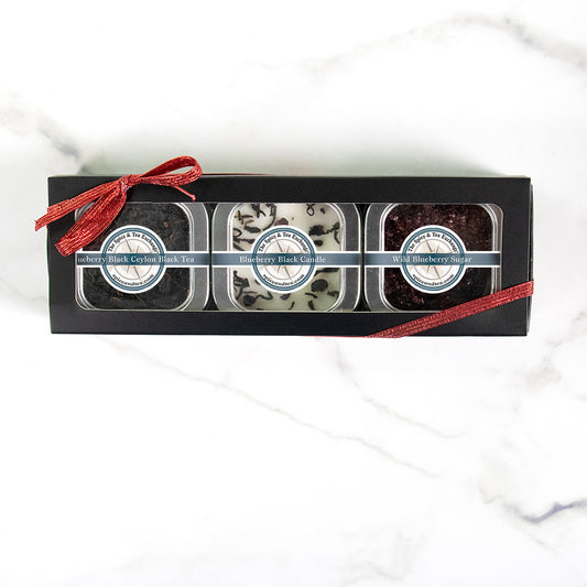 The Blueberry Black Tea Time Trio is a perfect tea gift set in a black box with a red ribbon, featuring Blueberry Black Tea, a matching candle, and Wild Blueberry Sugar—all beautifully labeled and displayed through a clear window.