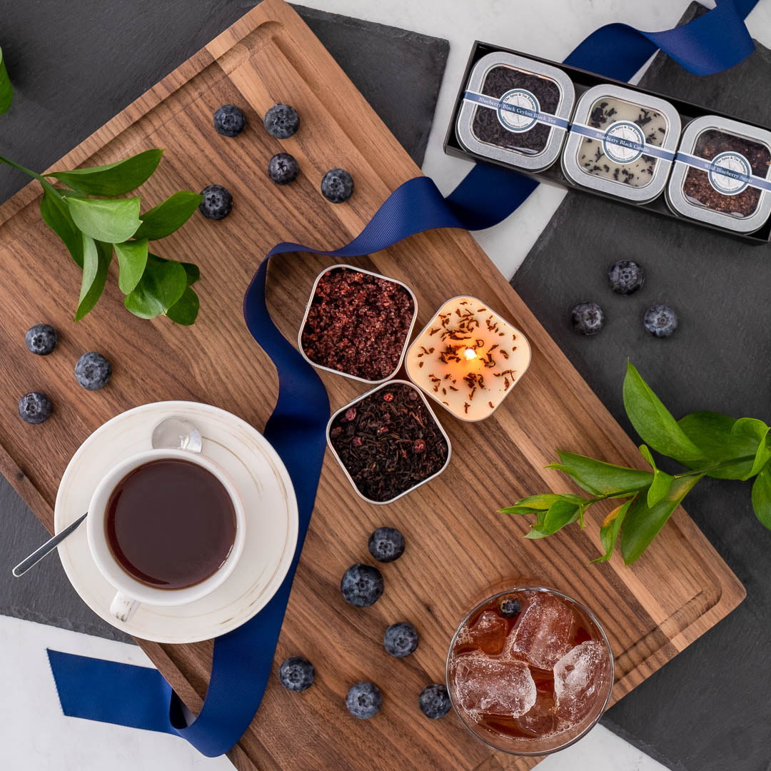 The Blueberry Black Tea Time Trio features a wooden tray with tea, an iced drink, loose tea leaves, a Blueberry Black Candle, blueberries, green leaves, a blue ribbon, and chocolates—an ideal gift for any tea lover.