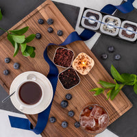 The Blueberry Black Tea Time Trio features a wooden tray with tea, an iced drink, loose tea leaves, a Blueberry Black Candle, blueberries, green leaves, a blue ribbon, and chocolates—an ideal gift for any tea lover.
