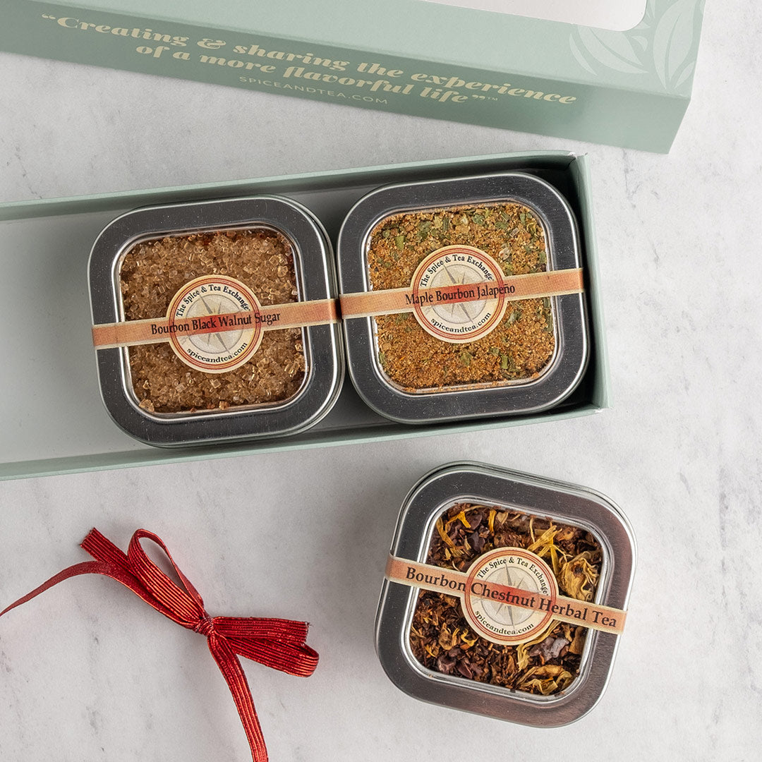 Three square metal tins of Bourbon herbal tea and spices arranged on a light marble surface, with one tin inside an open pale green gift box and another tied with a red ribbon.