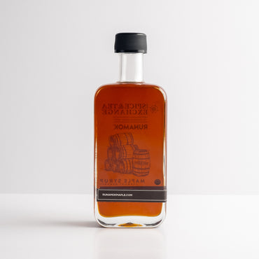 A rectangular glass bottle of Organic Barrel-Aged Bourbon Maple Syrup with dark amber syrup stands upright on a white surface. Its label features barrels and "pure Vermont maple syrup," finished with a black cap.