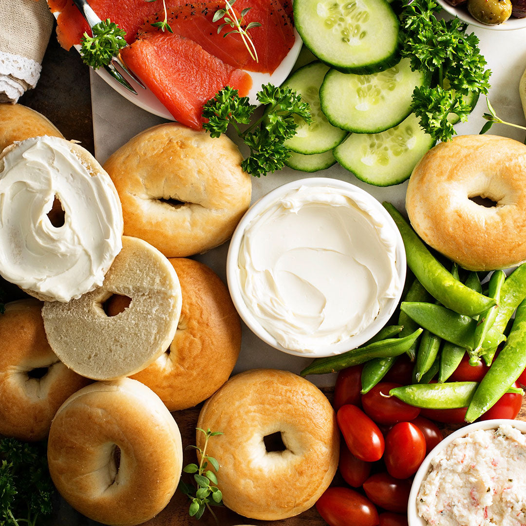 Platter with bagels, cream cheese, cucumbers, tomatoes, and snap peas on a wooden surface.