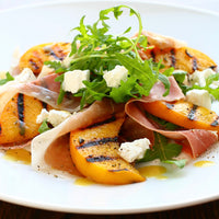 Salad with grilled peaches, prosciutto, and arugula on a white plate.