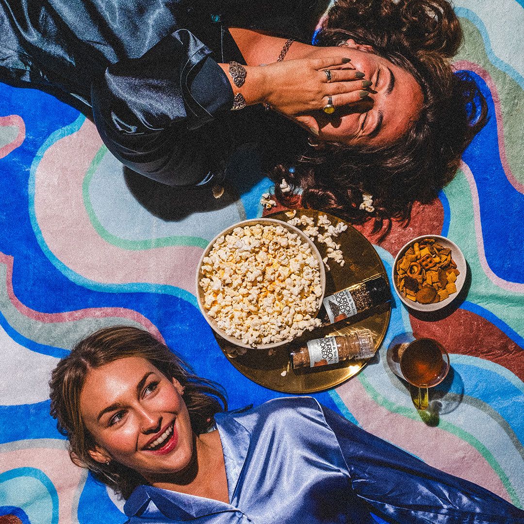 Two people lying on a colorful blanket with popcorn, drinks, and snacks.