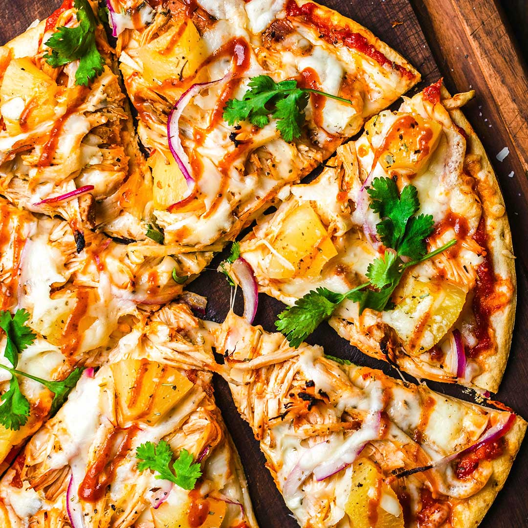 Pizza on wooden cutting board topped with grilled chicken, grilled pineapple, cilantro, red onion slices