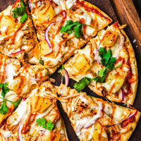 Pizza on wooden cutting board topped with grilled chicken, grilled pineapple, cilantro, red onion slices