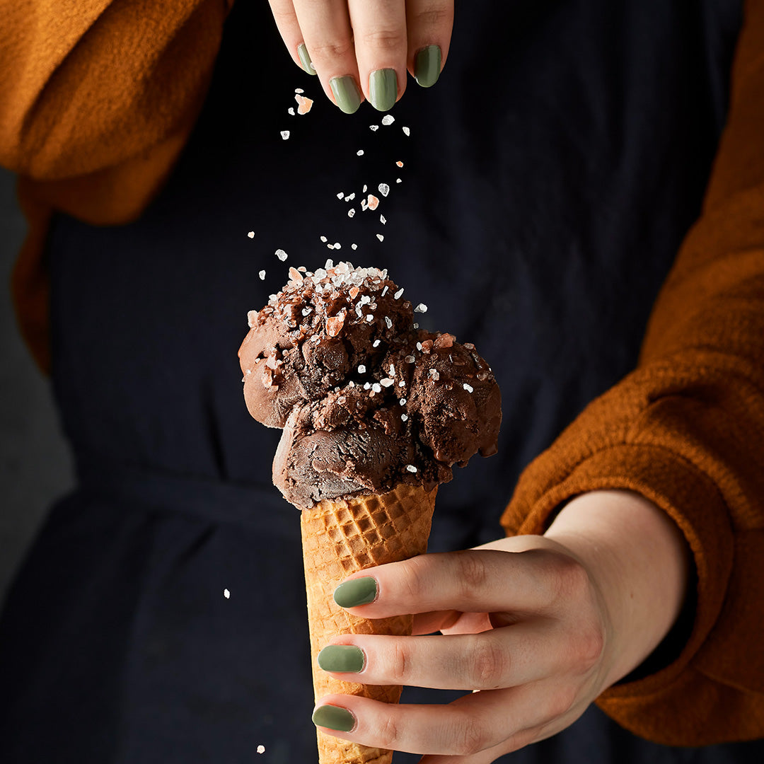 Chocolate ice cream cone being sprinkled with toppings by a person wearing an orange sweater.