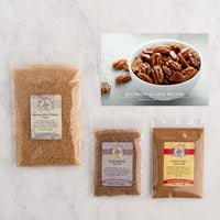 Three clear plastic bags of spices and ingredients arranged on a white marble surface, with a small recipe card showing bourbon glazed pecans in a bowl.