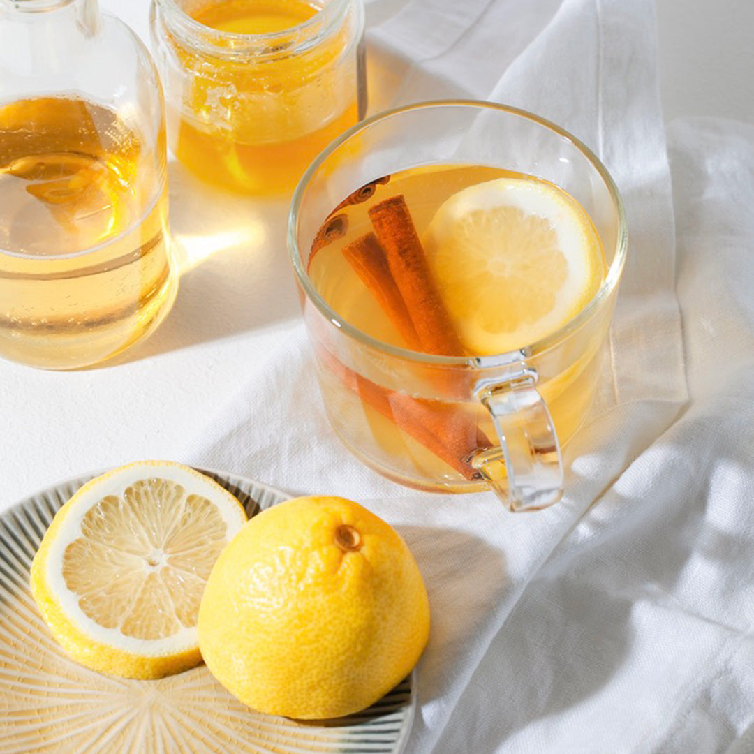 Clear mug with hot beverage, lemon slices, and cinnamon sticks on a white cloth background