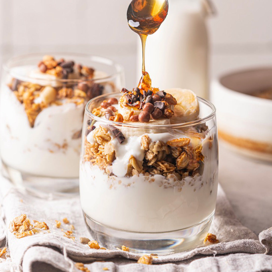 Glass of yogurt with granola and banana, honey being drizzled on top.