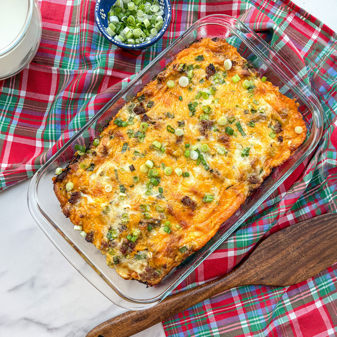 A baked casserole dish filled with melted cheese, green onions, and sausage, photographed from above on a red plaid cloth with a wooden spoon and a small bowl of chopped green onions.