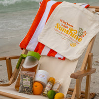 Bring Home A Little Sunshine Tote Bag hanging on beach chair next to Florida Sunshine products and orange and white beach towel with sand and ocean in background.