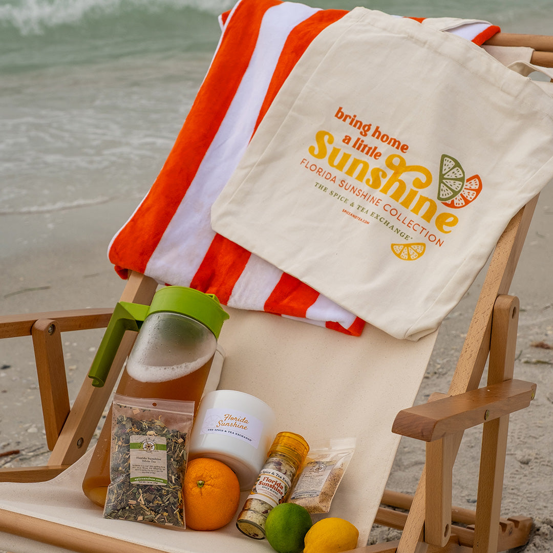 Bring Home A Little Sunshine Tote Bag hanging on beach chair next to Florida Sunshine products and orange and white beach towel with sand and ocean in background.