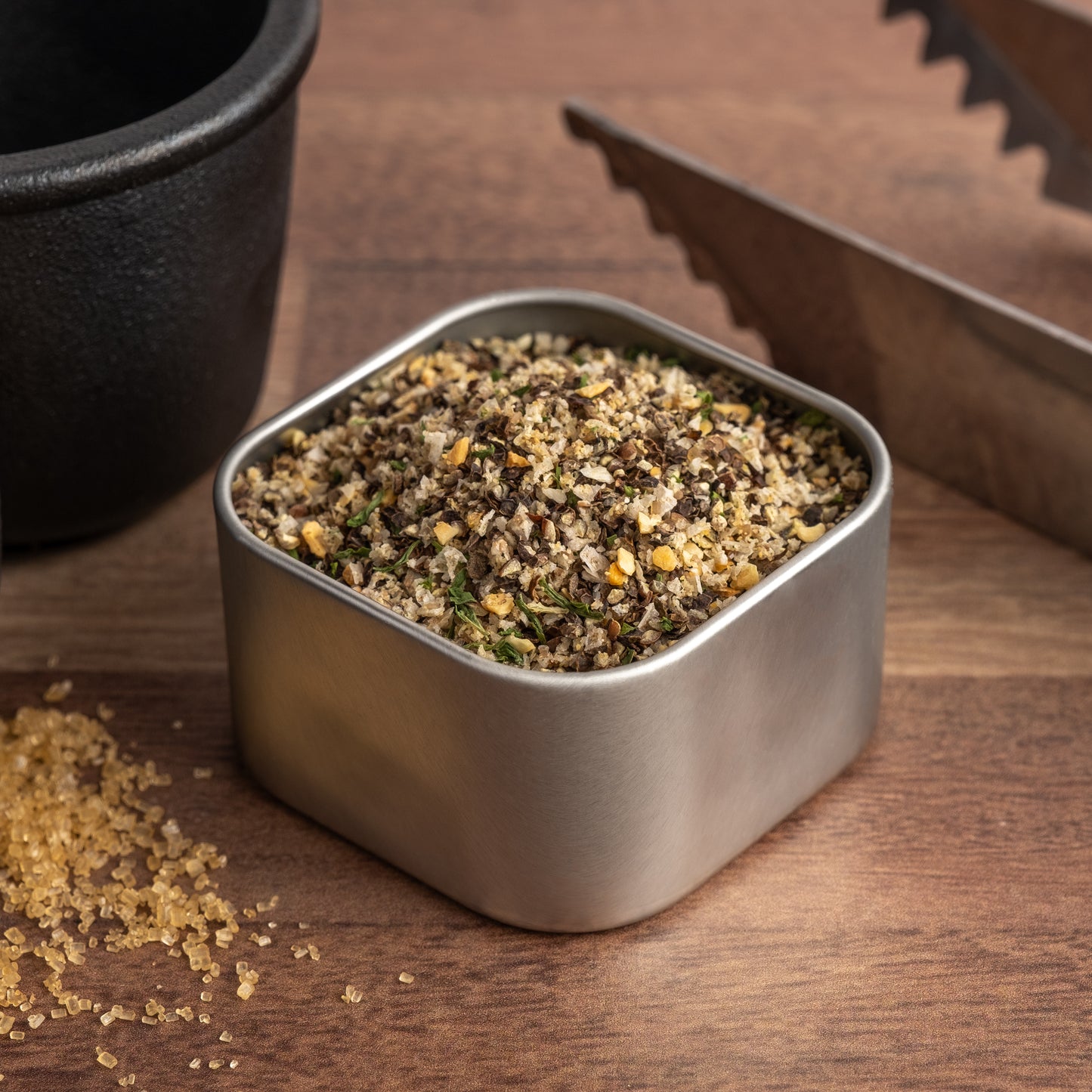 A square metal container of Butcher's Rub steak seasoning sits on a wooden surface beside tongs and a black bowl, with some coarse granules of the grilling spices scattered nearby.