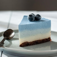 A slice of layered blue, white, and brown cheesecake on a light gray plate, topped with two fresh blueberries, with a silver spoon on a white table.
