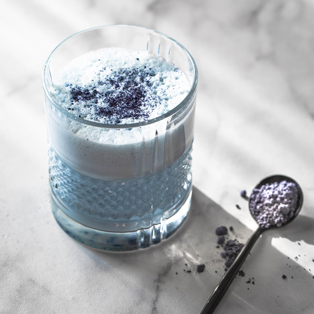 A textured clear glass filled with a light blue frothy liquid and topped with dark purple powder, set on a light marble surface with a spoon of the same purple powder beside it.