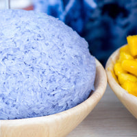 A close-up of a mound of light blue sticky rice in a light wood bowl, with a partially visible second bowl of yellow mango pieces at the right edge.