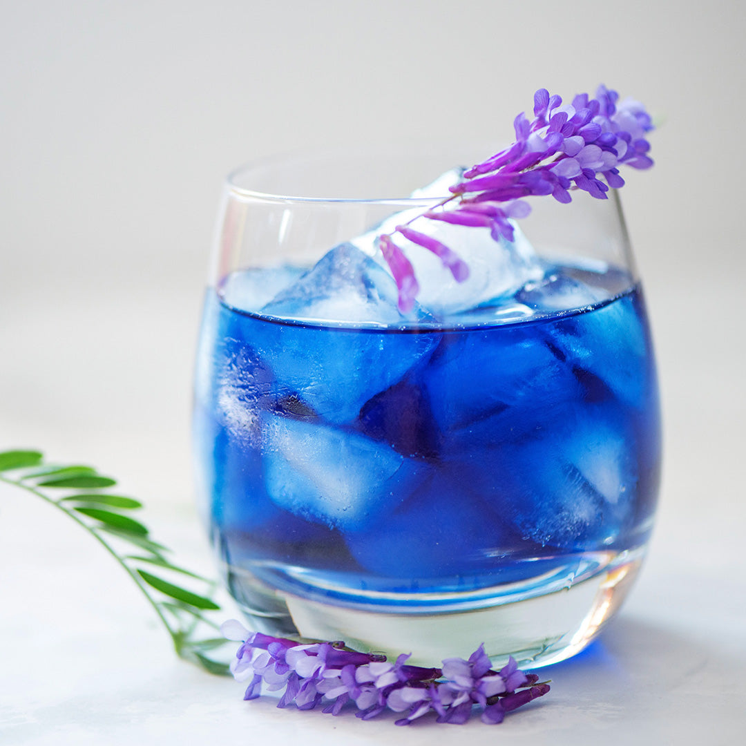 A clear stemless glass filled with vivid blue iced beverage and large ice cubes, garnished with purple flower sprigs, photographed on a clean light background.
