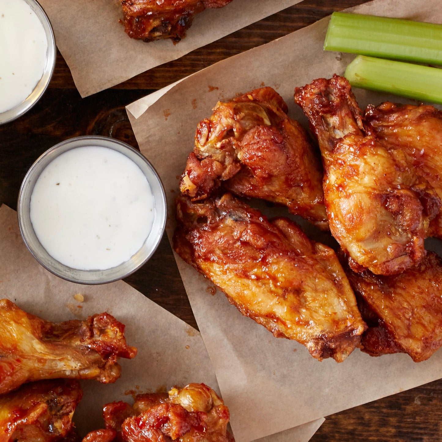 Crispy buffalo-style chicken wings with a glossy red-orange sauce, served on brown paper with small bowls of white dipping sauce and celery sticks on a dark wooden table.