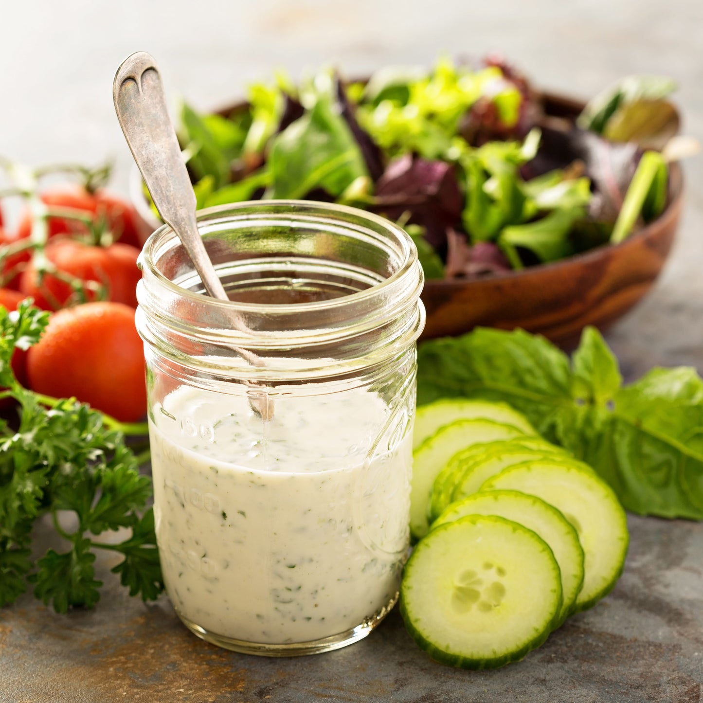 Jar of creamy dressing with a spoon, surrounded by fresh vegetables on a stone surface.