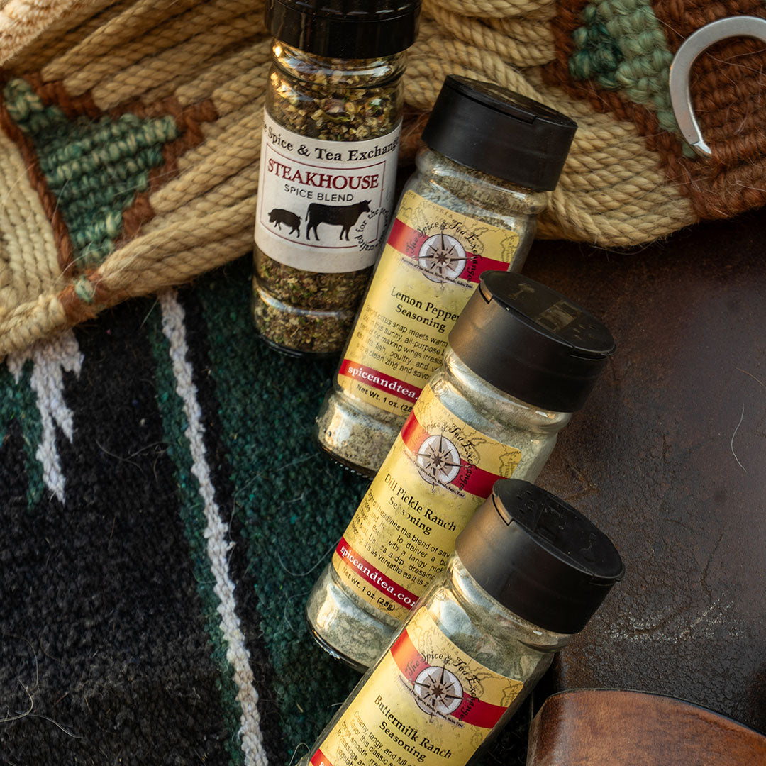 Four spice bottles from Spice & Tea Exchange arranged on a dark textured surface with a woven rug in the background. The bottles include Steakhouse Seasoning, Lemon Pepper Seasoning, Dill Pickle Ranch Seasoning, and Buttermilk Ranch Seasoning