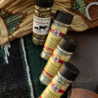 Four spice bottles from Spice & Tea Exchange arranged on a dark textured surface with a woven rug in the background. The bottles include Steakhouse Seasoning, Lemon Pepper Seasoning, Dill Pickle Ranch Seasoning, and Buttermilk Ranch Seasoning