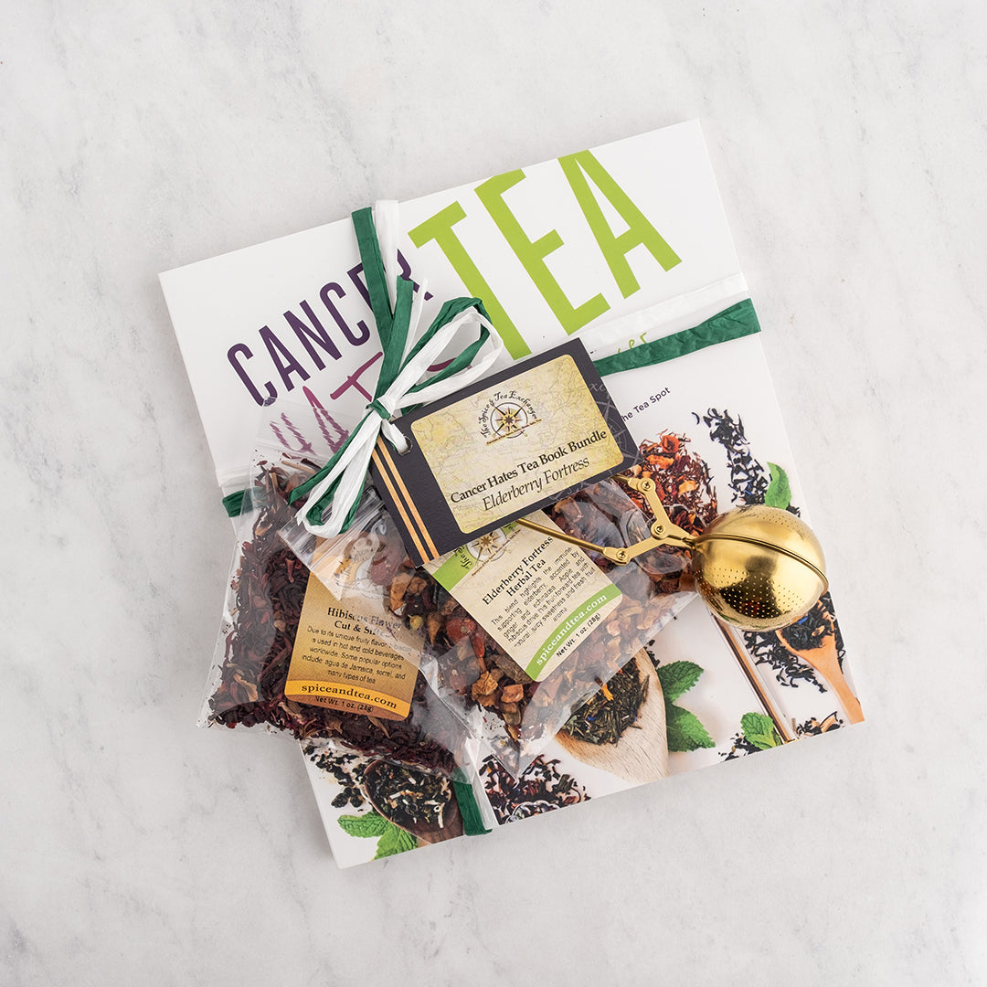 A tea gift set arranged on a white marble surface, featuring a white book, Elderberry Fortress Herbal Tea, Hibiscus Flower - Cut & Sifted, a green ribbon, a small gold scissors infuser, and a tag reading 'Cancer Home Tea Book Bundle Elderberry Fortress.'