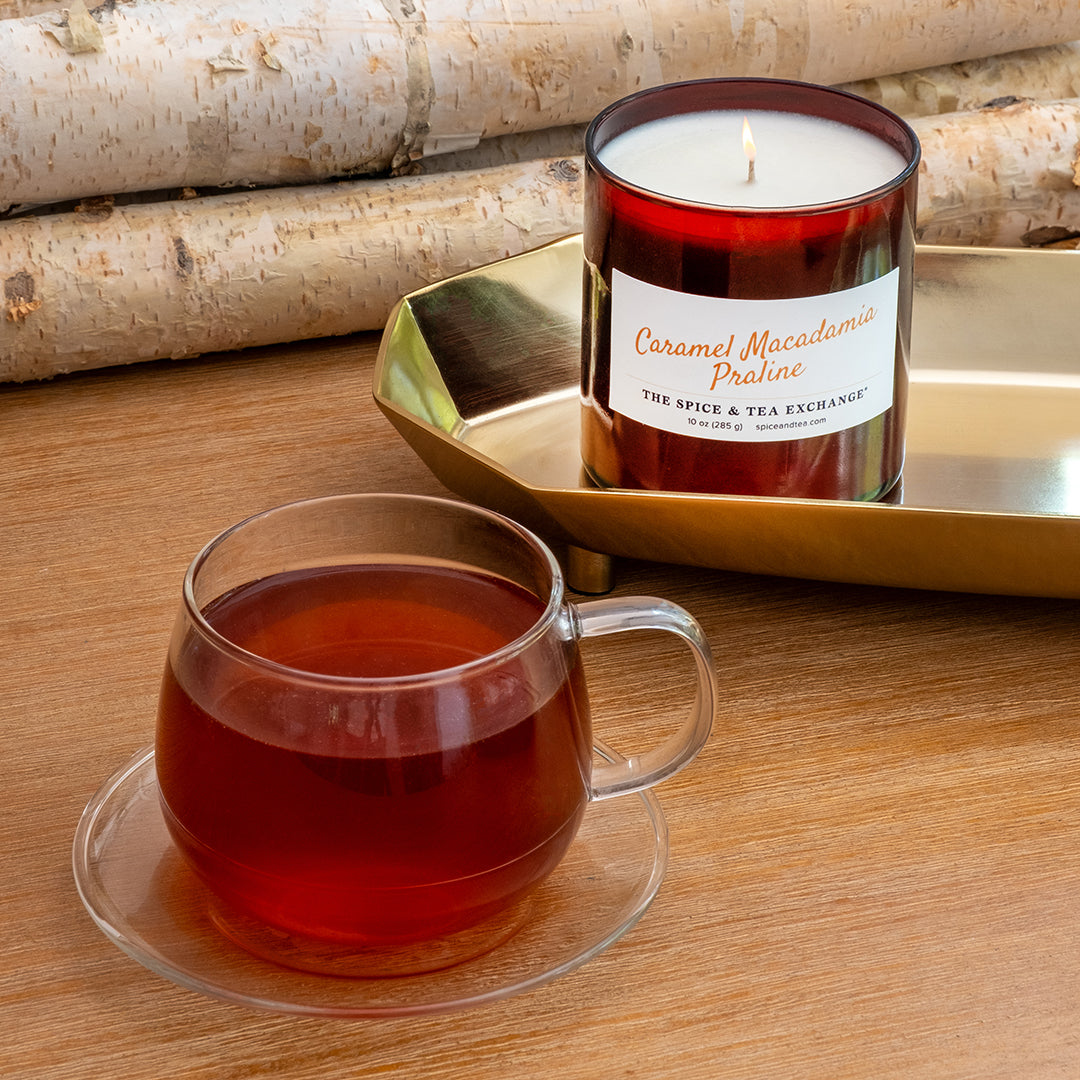 A glass jar candle labeled 'Caramel Macadamia Praline' sits on a gold tray beside a clear glass mug filled with amber tea on a wooden table, with birch logs in the background.