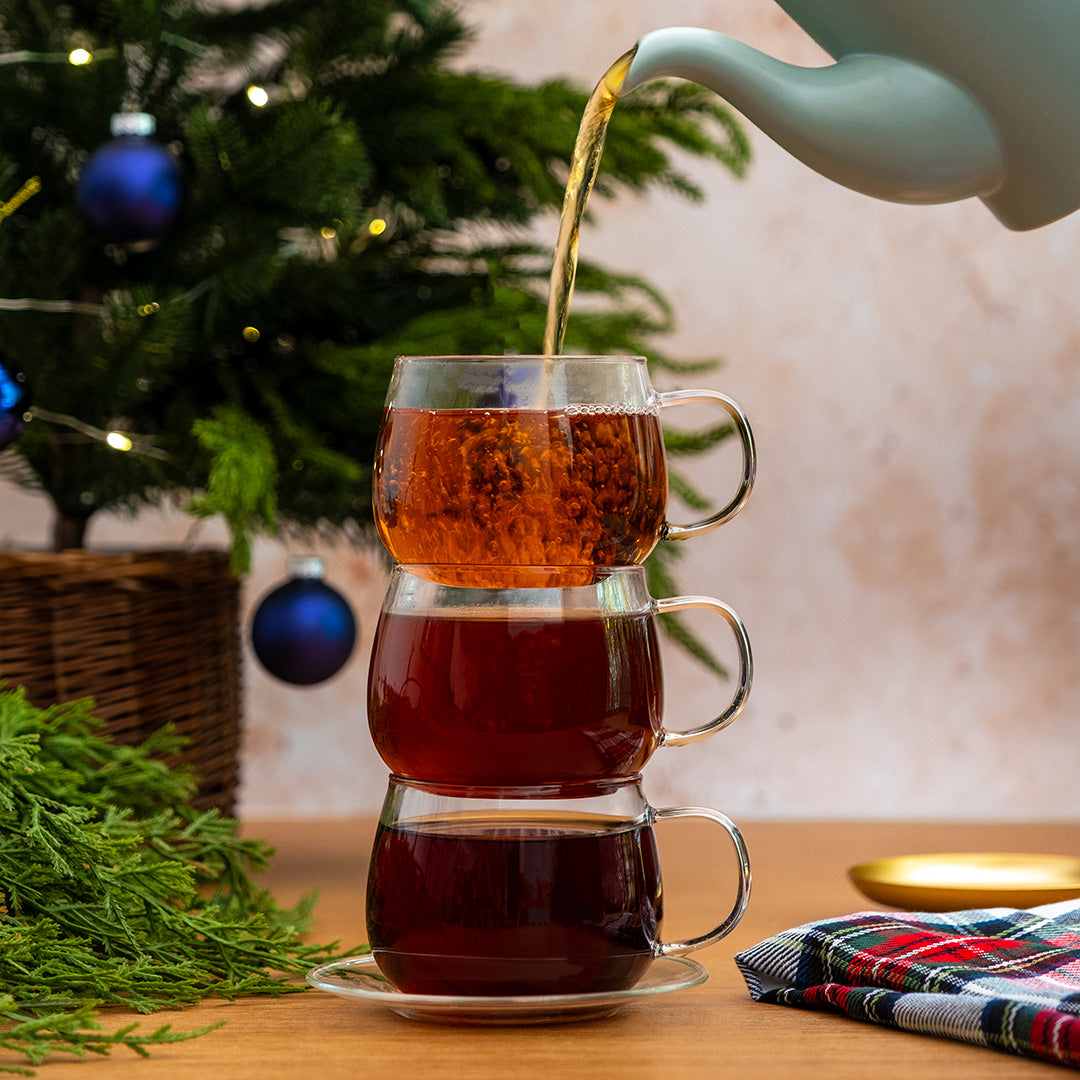 Three stacked clear glass teacups are filled with tea, with hot tea being poured from a white teapot. The cups sit on a wooden table with a Christmas tree, greenery, and a plaid cloth in the background.