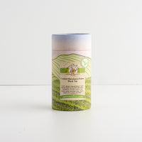A cylindrical cardboard tube package for Caramel Macadamia Praline Black Tea, featuring a scenic green hill illustration on a white background.