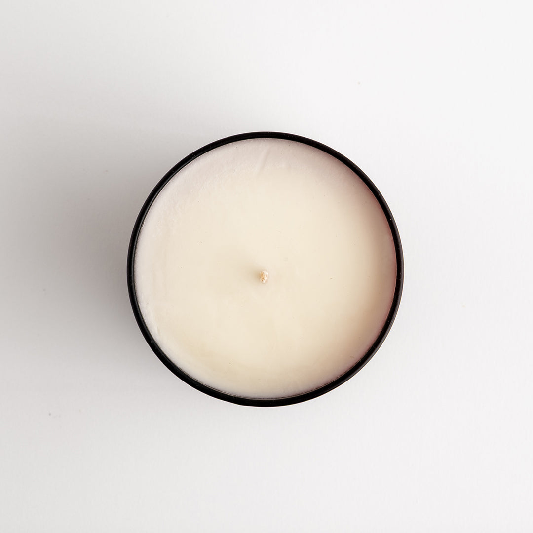A top-down view of a single-wick candle with creamy off-white wax inside a amber glass tin, set against a plain white background.