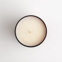 A top-down view of a single-wick candle with creamy off-white wax inside a amber glass tin, set against a plain white background.