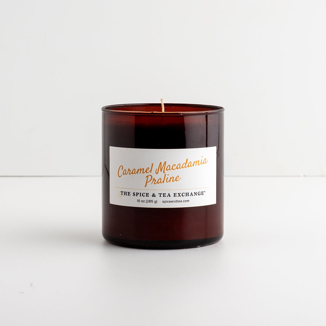 An amber glass jar candle with a white label reading 'Caramel Macadamia Praline' and 'THE SPICE & TEA EXCHANGE', photographed on a clean white surface against a white background.