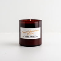 An amber glass jar candle with a white label reading 'Caramel Macadamia Praline' and 'THE SPICE & TEA EXCHANGE', photographed on a clean white surface against a white background.