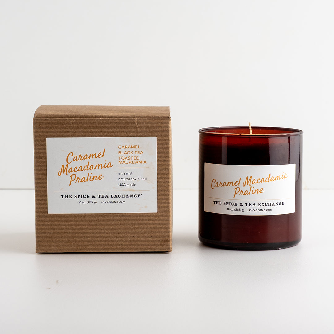 A candle in an amber glass jar next to its brown kraft paper box, both featuring white labels with orange text for 'Caramel Macadamia Praline' by 'The Spice & Tea Exchange'.