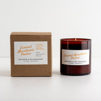 A candle in an amber glass jar next to its brown kraft paper box, both featuring white labels with orange text for 'Caramel Macadamia Praline' by 'The Spice & Tea Exchange'.