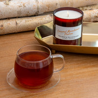A candle in a dark amber glass jar labeled 'Caramel Macadamia Praline' sits on a gold tray beside a clear glass mug filled with amber tea on a wooden table, with birch logs in the background.