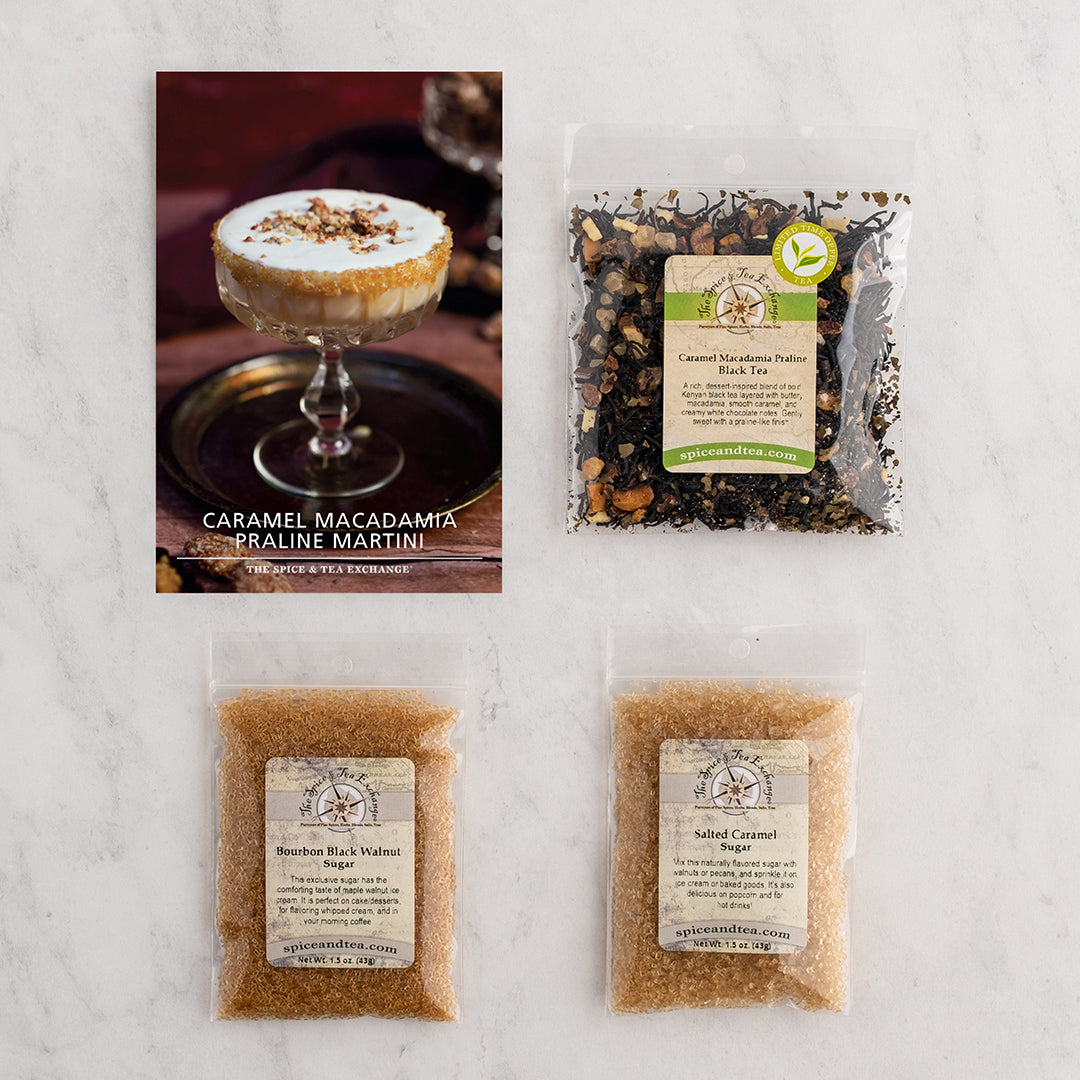 Four packaged food items arranged on a light marble surface: a recipe card for Caramel Macadamia Praline Martini, a bag of Caramel Macadamia Praline Black Tea, and two bags of sugar.