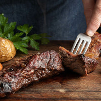 A person cuts into a juicy, grilled steak seasoned with Carne Asada (Kit) on a wooden board, with a baked potato and fresh parsley in the background.