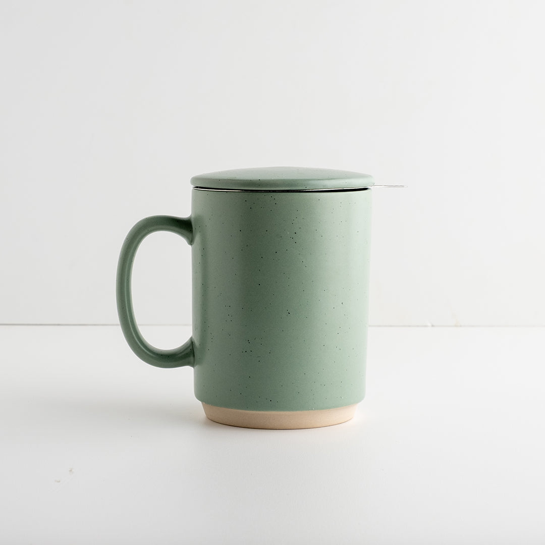 A speckled sage green ceramic mug with a matching lid and a small metal spoon resting on the side, set against a clean white background.