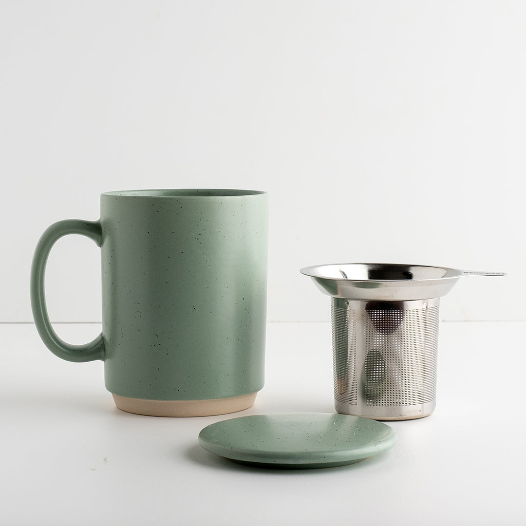 A sage green speckled ceramic mug with a matching ceramic lid, shown next to a stainless steel mesh tea infuser on a clean white background.
