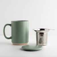 A sage green speckled ceramic mug with a matching ceramic lid, shown next to a stainless steel mesh tea infuser on a clean white background.